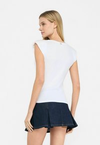 Top bianco senza maniche con un design aderente e sottili spalline, caratterizzato da un piccolo accento dorato sul retro, abbinato a una gonna di denim scuro.