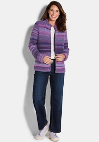 Frau lächelt, trägt eine lila gestreifte Strickjacke, ein weißes Shirt, dunkle Jeans und weiße Sneaker und steht vor einem schlichten Hintergrund.