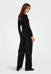 Svart langermet topp med ribbestruktur, kombinert med høytlivssvarte bukser med tynne striper. Glatt tekstur, skreddersydd passform og elegante svarte sko.