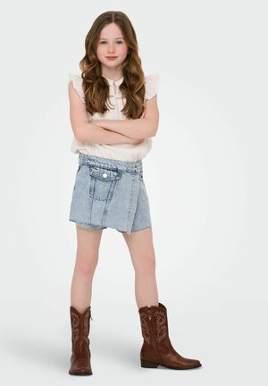 Jeansrok met rafelige zoom, gecombineerd met bruine cowboylaarzen met decoratieve stiksel. Witte blouse met ruches aan de mouwen en details bij de hals.