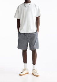 Biała bawełniana koszulka, szare szorty z elastycznym pasem, białe tenisówki z beżową podeszwą i widoczne białe skarpetki do kostek. Widoczny srebrny łańcuszek.