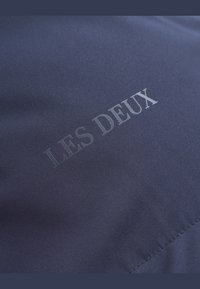 Marineblauer Stoff mit einer glatten Textur zeigt den eingeprägten Text "LES DEUX" in einem kontrastierenden Ton, der diagonal über die Oberfläche angeordnet ist.