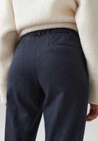 Pantaloni nautici scuri su misura con motivo a spina di pesce, cintura elastica e tasca posteriore. Abbinati a un maglione di pile color crema.