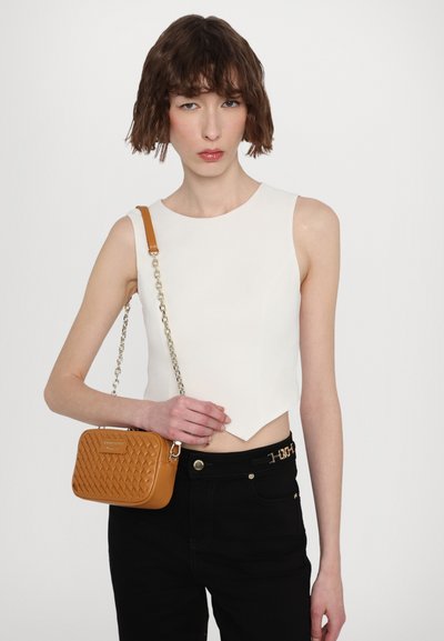 White sleeveless top, black pants, holding a small tan quilted handbag with a chain strap. Subtle design details visible on the garments.