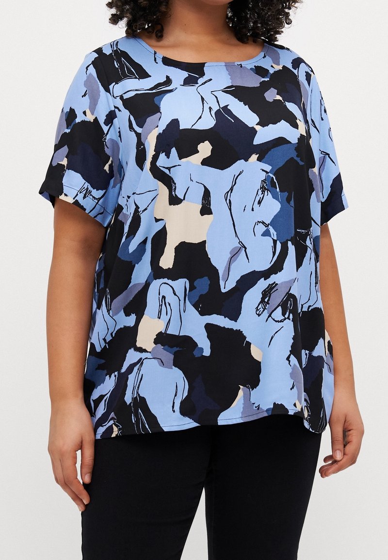 Femme portant un haut ample à manches courtes avec un motif abstrait bleu, noir, blanc et beige et un pantalon noir.