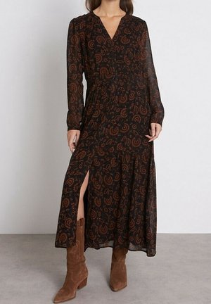 Femme portant une longue robe noire avec des motifs floraux marron, des manches longues, une fente à l'avant, associée à des bottes en daim marron mi-mollet.