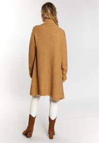 Suéter de punto color camel con cuello alto, mangas largas y dobladillo curvado, combinado con pantalones blancos y botas de tobillo marrones.