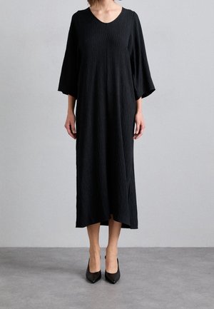 Kobieta w teksturowanej, czarnej sukience midi z rękawami do połowy przedramion i czarnych spiczastych szpilkach na szarej podłodze i tle.