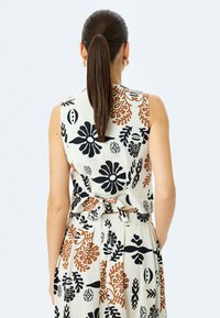 Vestido sin mangas en tejido crema con un patrón floral en negro y óxido, que presenta un detalle de lazo en la cintura y una falda plisada.
