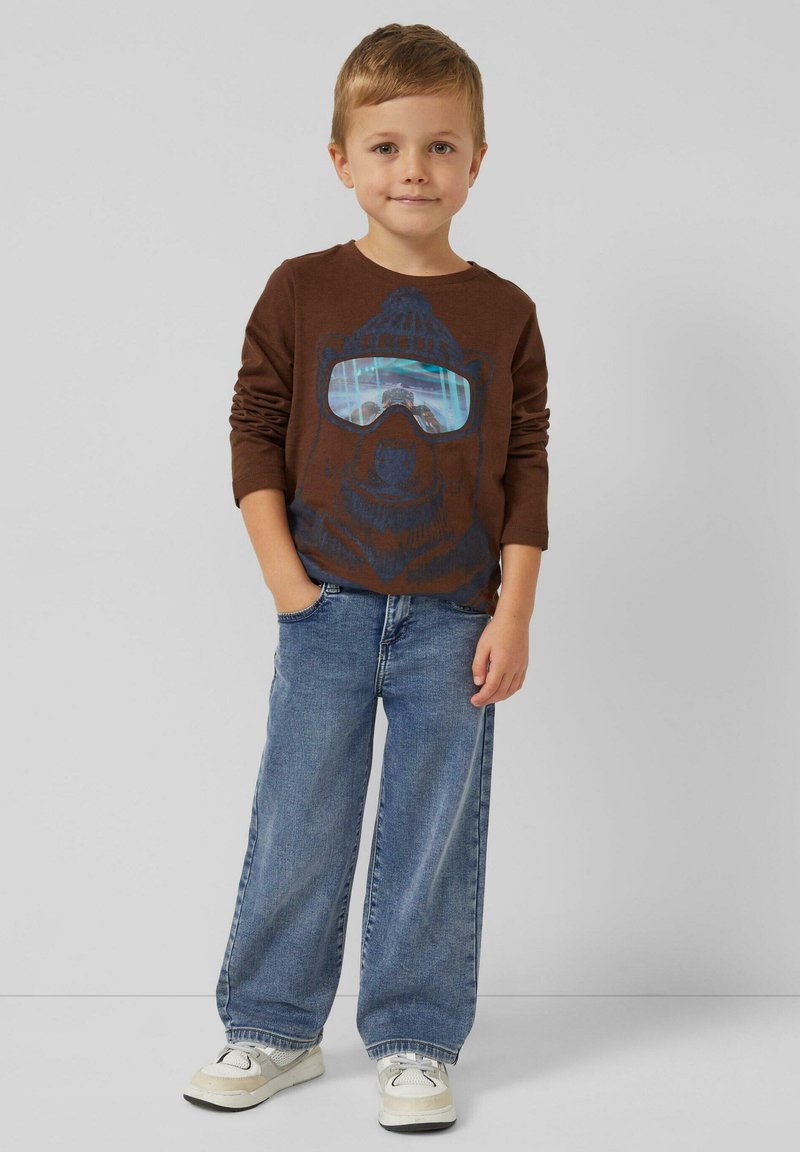 Braunes Langarmshirt mit einem Bär-Motiv, der eine Sonnenbrille trägt, kombiniert mit hellblauen, weitgeschnittenen Jeans und weißen Turnschuhen.