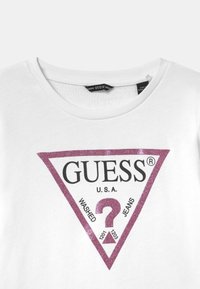 Camiseta de algodón blanca con un logo de triángulo rosa brillante que incluye el texto "GUESS U.S.A. WASHED JEANS" y un signo de interrogación en el centro.