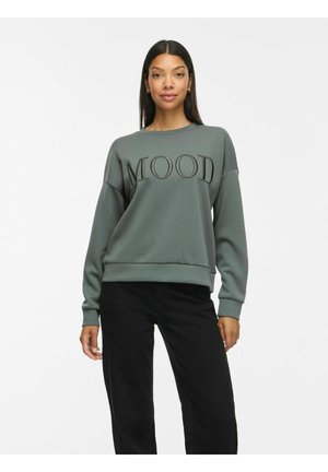 Femme aux longs cheveux foncés portant un sweat-shirt gris avec le mot "MOOD" en grandes lettres et un pantalon noir, debout sur fond blanc.