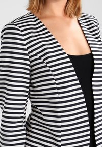 Svart och vit randig blazer med V-ringad design, med en figurnära passform och slät textur; öppen front utan knäppning.