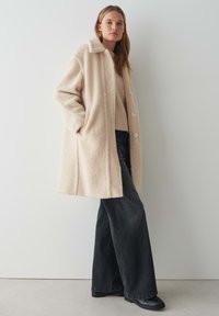 Cappotto oversize beige in finto shearling con colletto, chiusura frontale con bottoni e tasche laterali. Modello che indossa jeans svasati scuri e scarpe nere.