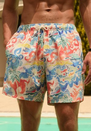 Taille und obere Beine eines oberkörperfreien Mannes, der mehrfarbige tropische Badeshorts mit Booten, Palmen und Meeresleben in der Nähe eines Pools trägt.