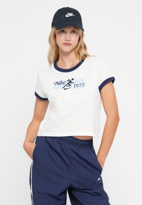 Balta īsa t-kreksa ar tumši zilu apdari, uz kuras ir drukāts Nike logotips un skrējēja grafika. Apvienota ar tumši ziliem biksēm.