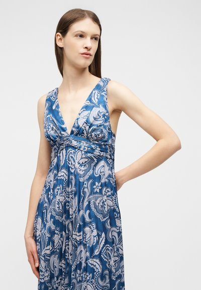 Jeune femme portant une robe bleue sans manches avec des motifs cachemire blancs, les mains positionnées à la taille et sur le côté, regardant sur le côté.