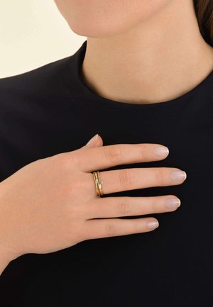 Goldfarbener Ring, der mit ineinandergreifenden Bändern und einem kleinen Diamantakzent verziert ist, wird an einem Finger getragen und zeigt eine glatte Oberfläche sowie ein zartes Design.