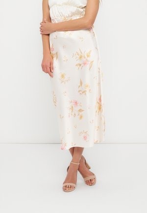 Ženska, oblečena v belo midi krilo z rožnatimi in bež cvetličnimi vzorci ter bež sandale z odprtom prstom in trakcem okoli gležnja, stoji pred belim ozadjem.