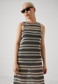 Robe en tricot présentant un motif rayé noir et crème, sans manches et avec une silhouette droite. Le modèle est accessoirisé avec des lunettes de soleil et des boucles d'oreilles.