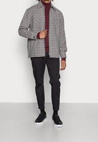 Chaqueta de camisa a cuadros en blanco y negro con dos bolsillos en el pecho, superpuesta a un cuello de tortuga color burdeos, combinada con pantalones negros y zapatillas negras.