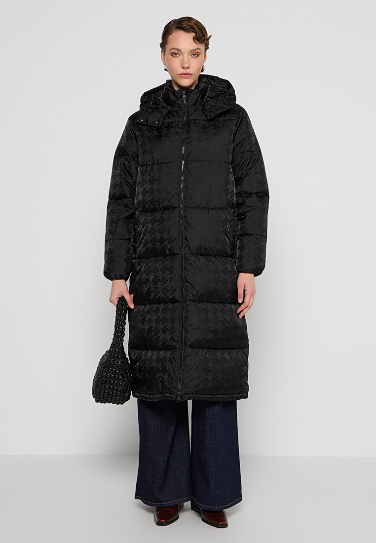 Schwarze Daunenjacke mit Kapuze und gestepptem Design. Das Modell trägt eine strukturierte schwarze Handtasche und weite, dunkle Hosen.