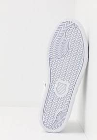 White rubber shoe sole featuring a herringbone traction pattern, rounded shape, and a shield emblem in the center.