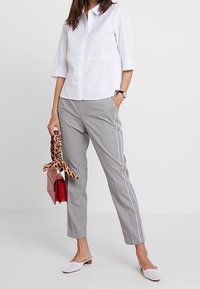 Vit skjorta med knappar, grå rutiga byxor med sidopiping, vita slip-on skor, röd handväska med ett mönstrat scarf som accent.