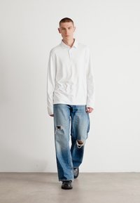 Hvit langermet polo skjorte kombinert med løse, medium blå jeans med hull på knærne. Modellen har på seg svarte sko mot en nøytral bakgrunn.