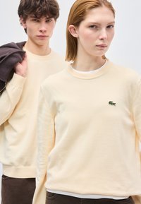 Homme et femme portant des pulls crème, la femme montrant un logo de crocodile vert sur son pull, l'homme tenant une veste sombre sur son épaule.