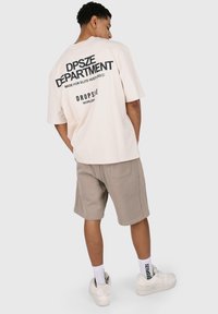 Kurze ärmeln, hellbeiges T-Shirt mit schwarzem Text auf dem Rücken, kombiniert mit beigen Shorts. Weiße Sneakers und Socken mit Markentext sind sichtbar.