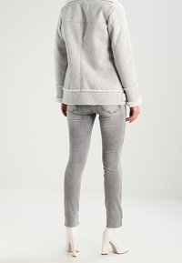 Veste en mélange de laine grise avec col en shearling, associée à un jean slim gris clair et des bottes à talons blanches, vue de dos.