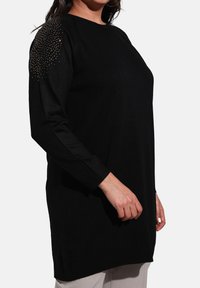 Black sweater with long sleeves, featuring a round neckline and decorative stud detailing on the shoulder. Lightweight knit fabric.