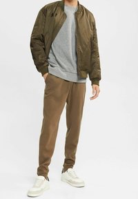 Giacca bomber verde oliva, felpa grigia sotto, pantaloni jogger marroni e sneakers bianche con dettagli crema. Vestibilità casual e rilassata.