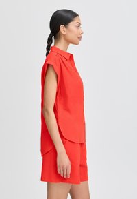 Camicia rossa a maniche corte con bottoni e orlo curvo, abbinata a pantaloni corti rossi coordinati, entrambi in un tessuto leggero.