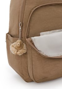 Mochila de tela marrón con un bolsillo con cremallera. Cuenta con un llavero de mono suave y esponjoso adjunto a la cremallera. El bolsillo delantero es de color crema.