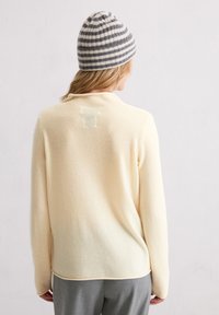 Kremowy sweter o miękkiej fakturze, z okrągłym dekoltem i subtelnym logo 'T' na plecach. Połączony z szarymi spodniami i paskowaną czapką.