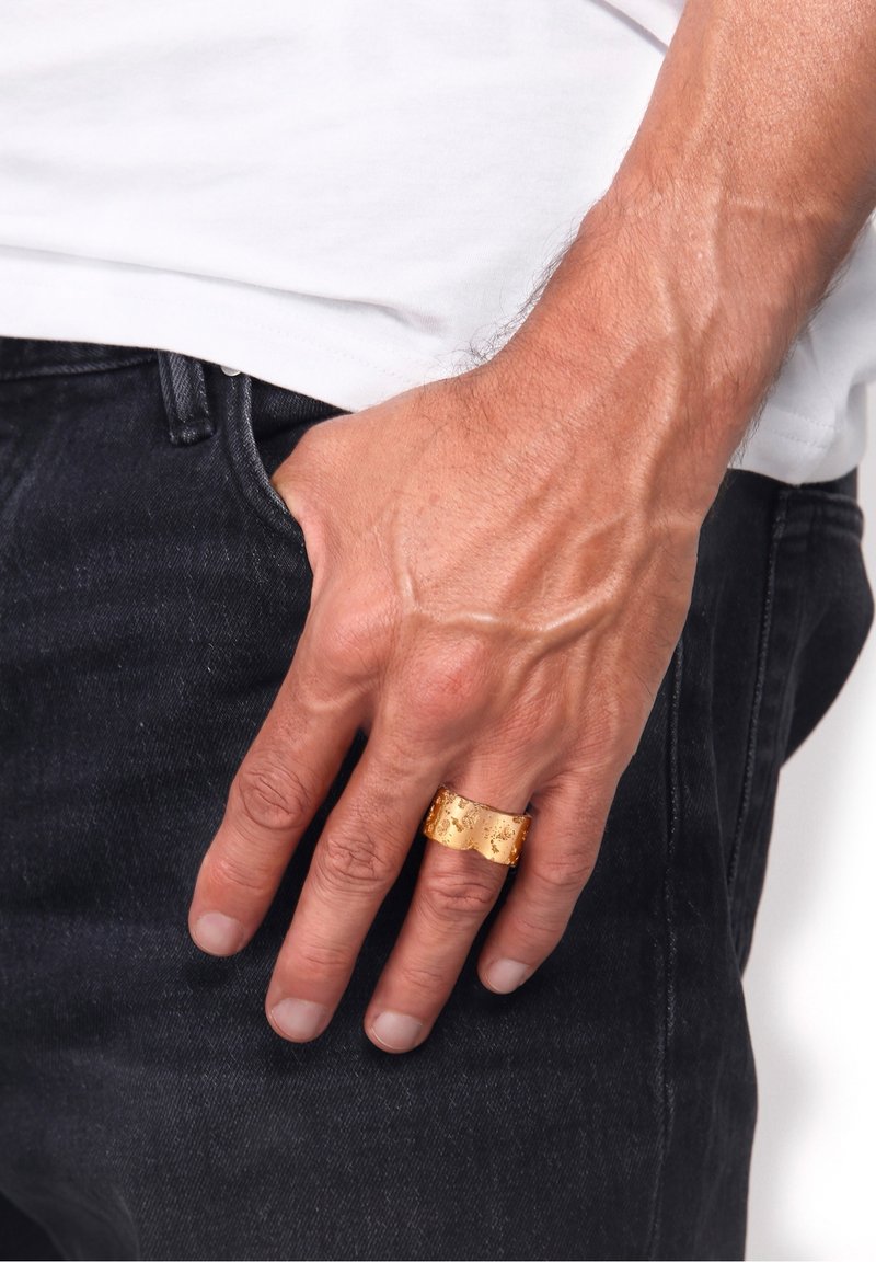 Main masculine avec des veines apparentes portant une large bague en or texturée sur le majeur, reposant près de la poche d'un jean noir.