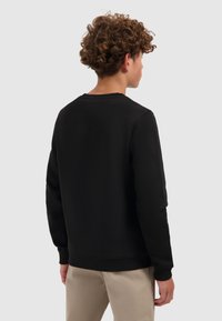 Jongere met krullend haar, gekleed in een zwarte longsleeve sweatshirt en beige broek, met de rug naar de camera, voor een effen lichte achtergrond.