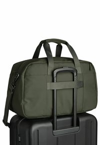 Sac polochon vert foncé posé sur la poignée déployée d'une valise noire rigide à roulettes.