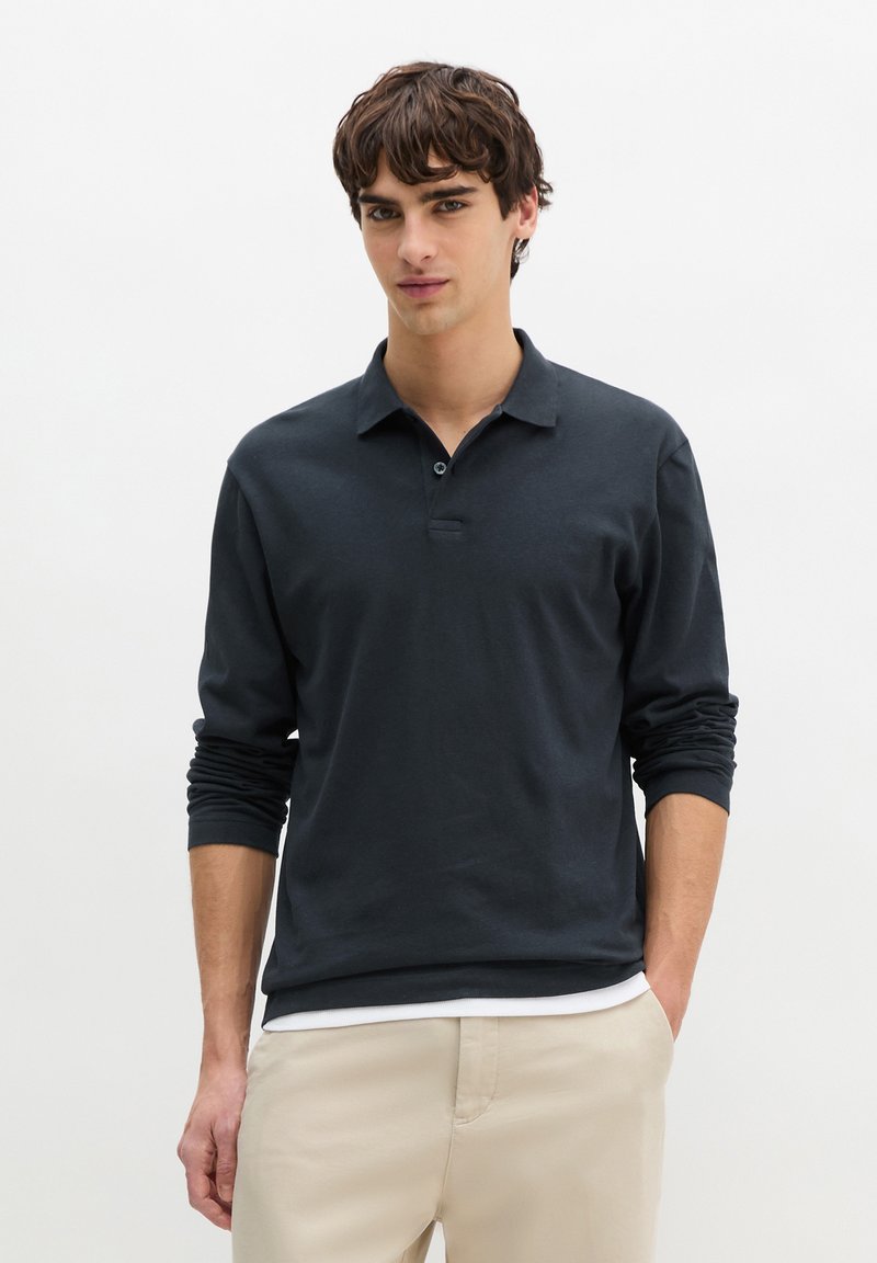 Jeune homme aux cheveux bruns ondulés, portant un polo noir à manches longues et un pantalon beige, debout les mains dans la poche.