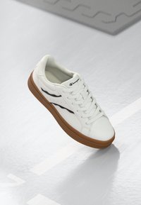 White athletic sneaker with a smooth leather upper, black accent stripes, and a tan rubber sole. Features flat laces and perforations for breathability.
