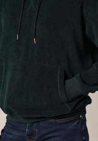 Haut à capuche en velours côtelé vert foncé avec un tissu texturé, des cordons de serrage noirs réglables et une poche frontale. Les poignets et l'ourlet sont côtelés.