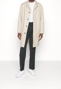 Mann trägt einen beigefarbenen, geknöpften Mantel, ein weißes Hemd, schwarze Hose und weiße Turnschuhe, steht vor einem weißen Hintergrund.