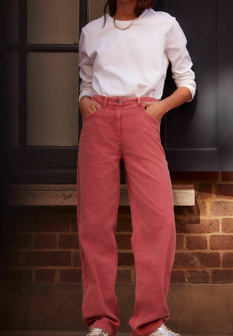Personne debout, mains dans les poches, portant une chemise blanche à manches longues, un collier en chaîne dorée, un pantalon en velours côtelé rouge et des baskets blanches.