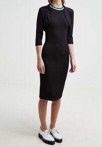 Svart figursydd klänning med trekvartslånga ärmar, med en rund halsringning och en grön och vit randig detalj. Matchas med vita skor.