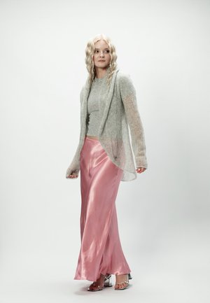 Kobieta o falowanych blond włosach, ubrana w szary dziany sweter, przezroczysty szary kardigan, długą różową satynową spódnicę oraz srebrne sandały na obcasie z odkrytymi palcami, stojąca na białym tle.