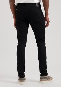 Jeans de mezclilla negra con un corte ajustado, bolsillos traseros y una etiqueta de la marca cosida en la cintura. Combinados con zapatillas gris y blancas.