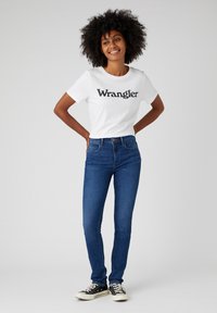 T-shirt bianca con logo nero "Wrangler", abbinata a jeans skinny blu e sneakers nere, con una vestibilità slim e uno stile classico.