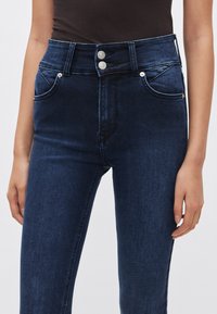 Jeans de mezclilla azul oscuro de talle alto, con cierre de dos botones, bolsillos delanteros y un sutil desgaste a lo largo de los muslos.